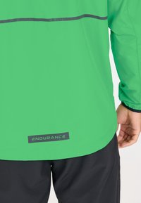 Veste légère de couleur vert vif avec une bande réfléchissante horizontale dans le dos. Étiquette noire "ENDURANCE" en bas. Tissu texturé et poignets élastiques.