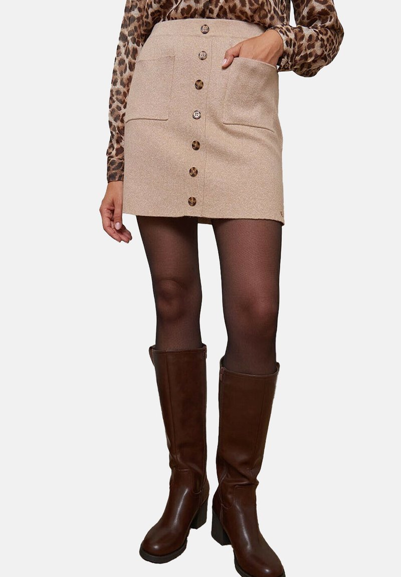 Jupe mini en tricot beige avec deux poches avant et fermeture à cinq boutons. Associée à des bottes en cuir marron à talons hauts.