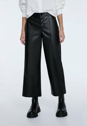 Persoon die zwarte wijde neplederen broek met voornaad draagt, zwarte enkellaarsjes met studs en een wit overhemd met lange mouwen die zijn opgerold.