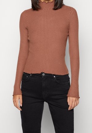 Pull en col montant côtelé marron avec un col festonné et des manches longues, associé à un jeans noir. Texture douce et coupe ajustée.