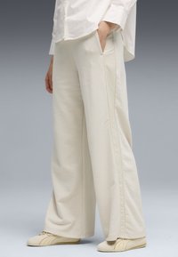 Personne portant un pantalon ample de couleur crème avec des poches latérales, des baskets Puma crème et une chemise blanche, debout devant un fond gris.