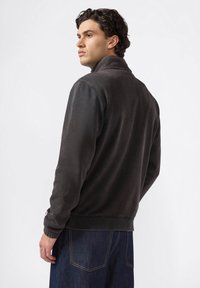 Joven con cabello rizado oscuro, vistiendo una chaqueta de forro polar gris oscuro y jeans azules, de perfil frente a un fondo blanco liso.