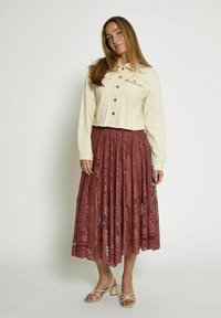 Giacca corta color crema con chiusura a bottoni, abbinata a una gonna maxi in pizzo bordeaux con intricate fantasie e texture fluida. Sandali beige con cinturini.