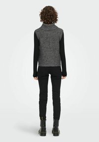 Personne avec des cheveux bouclés portant un gilet sans manches en tricot gris par-dessus un haut à manches longues noir, un pantalon noir et des bottes noires, se tenant de dos.