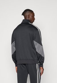 Μαύρο αθλητικό τζάκετ με ψηλό γιακά, γκρίζες πλευρικές πάνελ και λευκές ταινίες γύρω από τα μανίκια. Λείο υλικό, εφαρμοστή σχεδίαση.