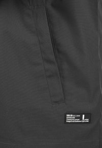 Veste noire avec une texture lisse et mate. Comprend une poche latérale et une étiquette affichant "SOLID EST.1999 Danemark" en texte blanc.