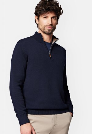 Granatowy sweter z dzianiny z zamkiem błyskawicznym przy kołnierzu typu mock neck, z prążkowanymi mankietami i dołem, z dyskretnym logo na piersi oraz gładką fakturą.