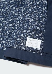 Tummansininen kangas, jossa vaaleansinainen ja kermanvärinen kukkakuvio. Mukana valkoinen etiketti, jossa on tilat nimelle ja puhelinnumerolle.