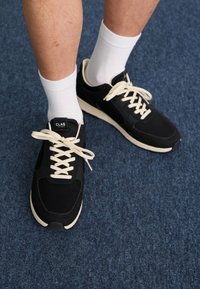 Sorte treningssko med beige detaljer og hvite lisser, brukt med hvite sokker, plassert på en blå teksturert overflate.
