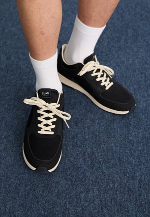 Mustat urheilukengät beige-aksentilla ja valkoisilla nauhoilla, joita pidetään valkoisten sukkien kanssa, sijoitettu siniselle teksturoidulle pinnalle.