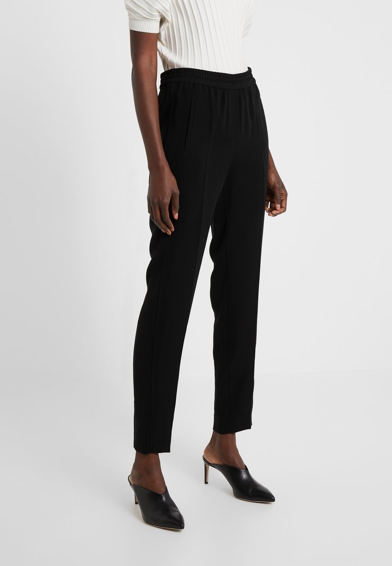 Personne portant un pantalon noir ajusté avec une taille élastique et des mules noires à talons aiguilles, accompagnés d'un haut à manches courtes blanc à côtes.