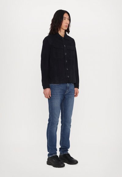 Veste noire à boutons avec deux poches poitrine, portée sur un jean bleu et des chaussures noires ; texture lisse et design structuré.