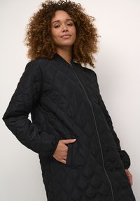 Veste noire matelassée avec une fermeture éclair à l'avant, un col ribbé, des poches latérales et un motif en losanges. Tissu lisse avec une silhouette ajustée.
