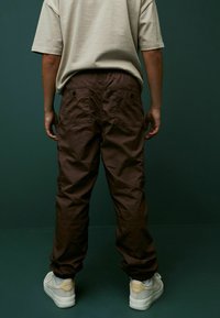 Braune Cargo-Hosen mit elastischen Bündchen, zwei Gesäßtaschen und einer lockeren Passform, kombiniert mit hellen Sneakern. Beiger T-Shirt von hinten sichtbar.