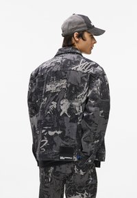 Μαύρο τζάκετ από τζιν με αφηρημένα γκρι γραφικά, κλείσιμο με κουμπιά και πλαϊνές τσέπες. Περιλαμβάνει ετικέτα μάρκας στο στρίφωμα και αντίθετα μπλε στοιχεία.