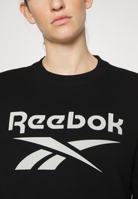 Felpa nera con collo a giro, dotata di un grande logo Reebok bianco e elementi di design grafico. Tessuto morbido con una texture liscia.