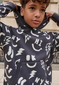 Dark gray hoodie featuring white graphic patterns of smiley faces, lightning bolts, and text. Cotton blend with a front pocket and ribbed cuffs.
