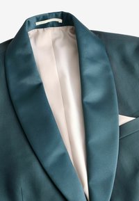 Blazer de satén en color verde azulado con cuello de pico, que presenta un forro suave de color claro y un pañuelo de bolsillo en un tono contrastante.