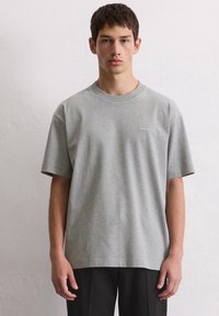 Młody mężczyzna ubrany w jednolitą szaro-skalną koszulkę Marc O'Polo i czarne spodnie, stojący na tle jasnoszarej ściany, zwrócony w kierunku frontu.