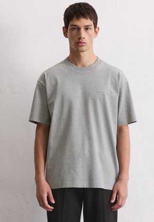 Young man wearing a plain grey Marc O'Polo t-shirt and black pants, standing against a light grey wall, facing front.