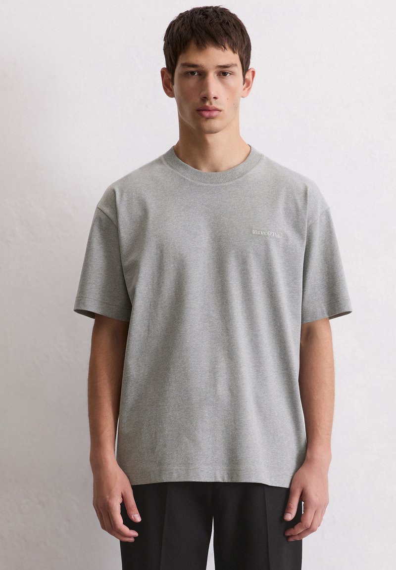 Młody mężczyzna ubrany w jednolitą szaro-skalną koszulkę Marc O'Polo i czarne spodnie, stojący na tle jasnoszarej ściany, zwrócony w kierunku frontu.