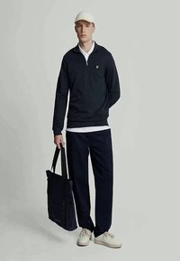Sweater azul-marinho com fecho, colarinho branco, combinado com calças largas azul-marinho e uma bolsa de mão correspondente; ténis creme completam o visual.