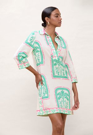 Mujer con vestido blanco hasta la rodilla con estampado tropical en verde y rosa, de pie de perfil con una mano en la cadera.