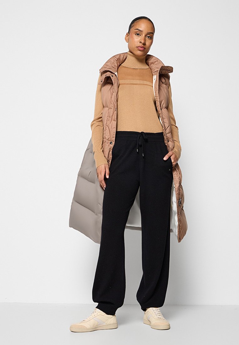 Braune Steppweste über einem beigen Sweater, kombiniert mit schwarzen Jogginghosen. Cremefarbene Sneakers runden das Outfit ab. Glatte Texturen, lässiges Design.