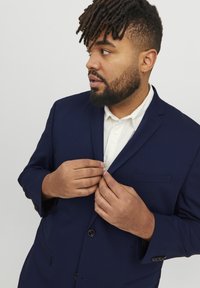 Dunkelblauer Blazer mit Reverskragen, zwei vorderen Knöpfen und einer Brusttasche; über einem weißen Hemd mit Kragen getragen. Strukturiert.