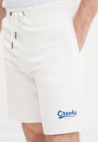 Witte katoenen shorts met een trekkoord taille, zijzakken en een blauw "Creeks" logo met een gele accent. Zachte textuur.
