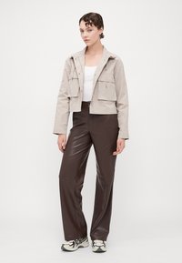 Beige cropped jacket with two front pockets, paired with brown high-waisted, shiny pants. Model wears white top and black and white sneakers.