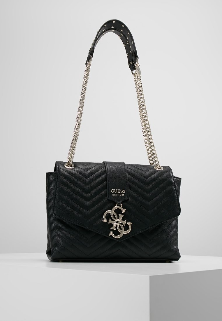 Sac à main Guess en cuir matelassé noir avec bandoulière chaîne dorée et breloque logo sur un socle d'exposition blanc.