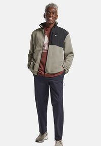 Veste en polaire grise avec une partie supérieure noire, associée à un t-shirt graphique bordeaux et un pantalon bleu marine. Les chaussures sont beige avec une finition texturée.