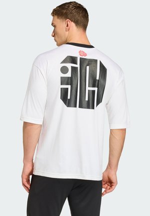 Homme de profil portant un T-shirt blanc surdimensionné avec un grand motif géométrique noir et une petite rose rouge sur le haut du dos.