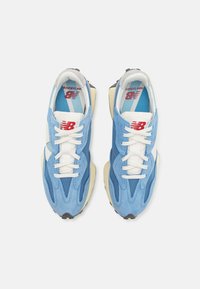 Modré semišové a síťované tenisky s bílými tkaničkami, které mají vrstvený design a kontrastní černou gumovou podešev. Červené logo na jazyku.