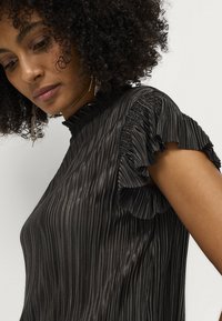 Sort plisseret bluse med høj hals og rynkede korte ærmer. Stoffet har en glat, blank overflade og et lodret stribet mønster.