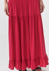 Lange rode gelaagde rok met een gerimpelde zoom, gedragen met zwarte sandalen, handen ontspannen langs de zijden tegen een effen achtergrond.