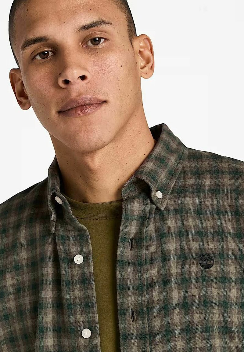 Jeune homme portant une chemise à carreaux verte et grise par-dessus un t-shirt olive sur fond uni.