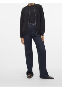 Blouson aviateur noir, fermeture éclair courte, deux poches ; associé à un jean bleu taille haute, jambes larges, tissu foncé, et chaussures plates noires.