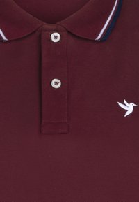 Camisa polo burdeos con un tejido de punto texturizado, una tapeta de tres botones y un logo de ave bordado en blanco en el lado izquierdo del pecho.