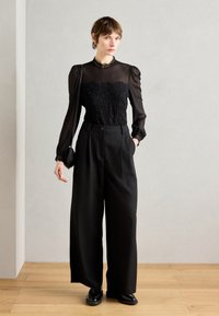 Zwarte transparante blouse met lange mouwen en kantdetails, gecombineerd met hoge taille wijde zwarte broek en zwarte schoenen. Eenvoudige handtas.
