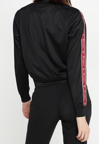 Svart atletisk jacka med långa ärmar, slät tyg, ribbade ärmslut och ett röd och vitt logoband längs ärmarna.