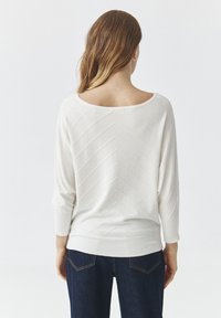 Hvid langærmet sweater med chevron struktur, bred halsudskæring og ribbede manchetter og kant. Båret med mørkeblå jeans.