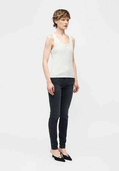 Femme debout portant un débardeur blanc à encolure dégagée, un jean skinny noir et des escarpins slingback noirs à bout pointu sur un fond uni.