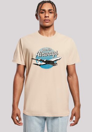 Mann trägt ein beiges T-Shirt mit dem Vintage-Logo der Pan Am Airways und einem Flugzeugmotiv, kombiniert mit hellblauen Jeans, neutraler Hintergrund.