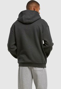 Felpa con cappuccio di color grigio scuro, dal taglio rilassato e realizzata in tessuto testurizzato. Presenta un ampio cappuccio e polsini a coste, abbinata a pantaloni della tuta grigi chiari.