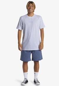 Camiseta de algodón blanca con logo azul, corte relajado, combinada con shorts azules, calcetines blancos de caña baja y zapatillas negras. Diseño simple, estilo casual.
