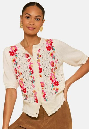 Žena v krémovom krátkom pletenom kardigáne s krátkym rukávom, ozdobenom ružovým a červeným kvetinovým výšivkom a ozdobnými vlnitými okrajmi, v kombinácii s hnedými nohavicami.