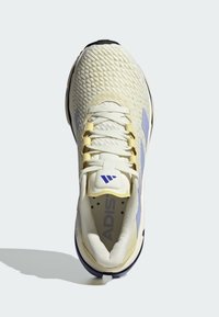 Scarpa sportiva con una tomaia in crema testurizzata, dettagli giallo pallido e suola blu navy. Presenta un colletto imbottito e un sistema di allacciatura evidente.
