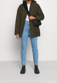 Parka verde oliva, jeans blu a vita alta, stivali neri alla caviglia. Posizione in piedi, borsa a tracolla nera minimalista, texture del tessuto liscia.